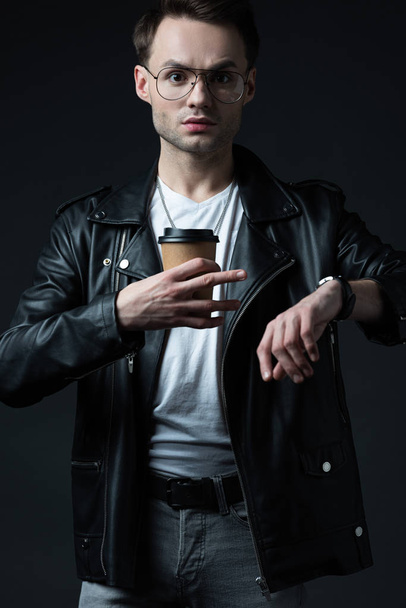 stressed stylish brutal man in biker jacket with wristwatch and coffee to go isolated on black - Fotografie, Obrázek