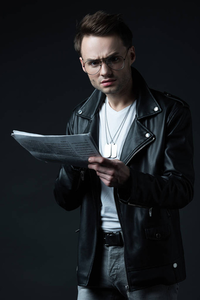 confused stylish brutal man in biker jacket with newspaper isolated on black - Foto, Imagen