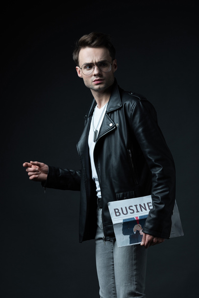 serious stylish brutal man in biker jacket with business newspaper isolated on black - Zdjęcie, obraz