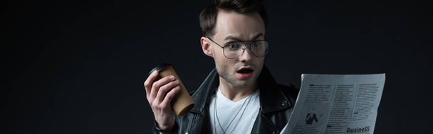 shocked stylish brutal man in biker jacket with newspaper and coffee to go isolated on black, panoramic shot - Photo, image