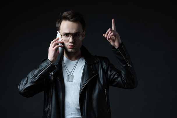 tense stylish man in leather jacket talking on smartphone and showing idea gesture isolated on black - 写真・画像