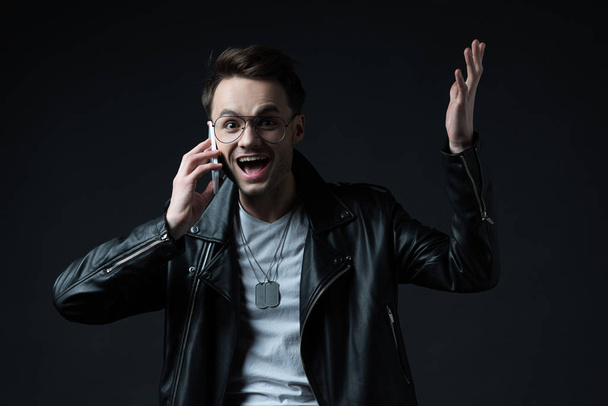 happy excited stylish man in leather jacket talking on smartphone isolated on black - Foto, imagen