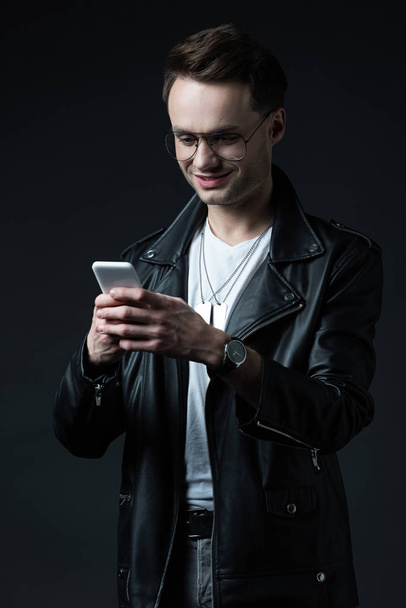 sorrindo homem elegante em jaqueta de couro usando smartphone isolado em preto
 - Foto, Imagem