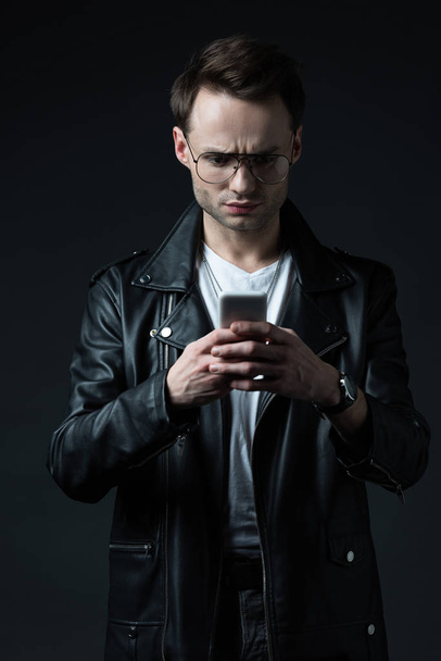 ανήσυχος κομψό άνδρα με δερμάτινο μπουφάν χρησιμοποιώντας smartphone απομονωμένο σε μαύρο - Φωτογραφία, εικόνα