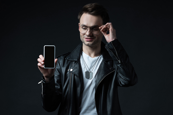 usmívající se stylový muž v kožené bundě prezentující smartphone s prázdnou obrazovkou izolované na černé - Fotografie, Obrázek
