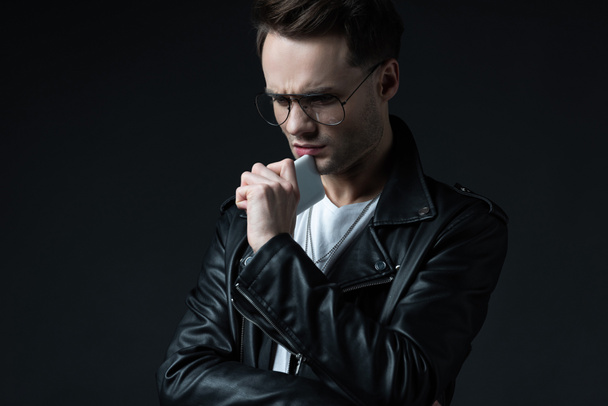 worried stylish man in leather jacket with smartphone isolated on black - Фото, изображение