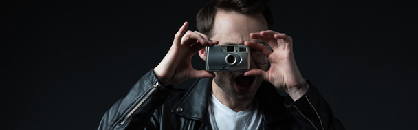 stylish brutal man in biker jacket taking picture on film camera with open mouth isolated on black, panoramic shot - Photo, Image
