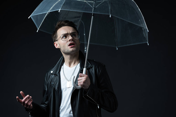 shocked stylish brutal man in biker jacket looking at umbrella isolated on black - Photo, Image