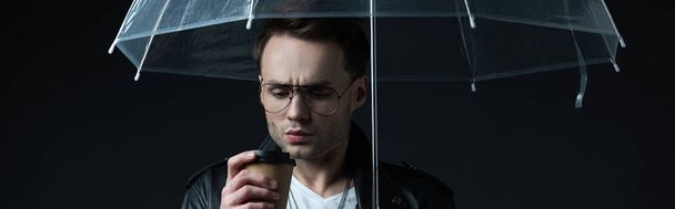 worried stylish brutal man in biker jacket with umbrella and coffee to go isolated on black, panoramic shot - Foto, imagen