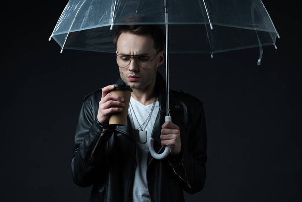 worried stylish brutal man in biker jacket with umbrella and coffee to go isolated on black - Photo, image