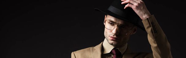 elegant man in beige suit and eyeglasses putting on hat isolated on black, panoramic shot - Foto, Imagen