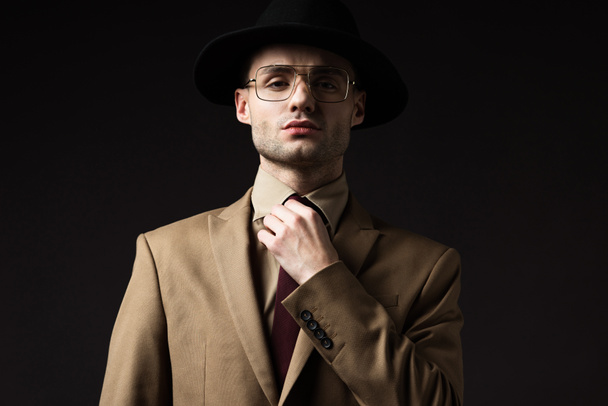 elegante man in beige pak, hoed en bril bevestigingsstropdas geïsoleerd op zwart - Foto, afbeelding