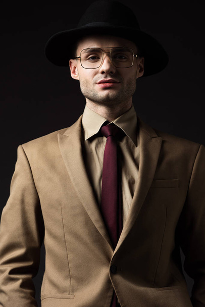 confident elegant man in beige suit, hat and eyeglasses isolated on black - Фото, зображення