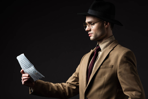 vista lateral del hombre elegante en traje beige, sombrero y anteojos leyendo periódico aislado en negro
 - Foto, Imagen