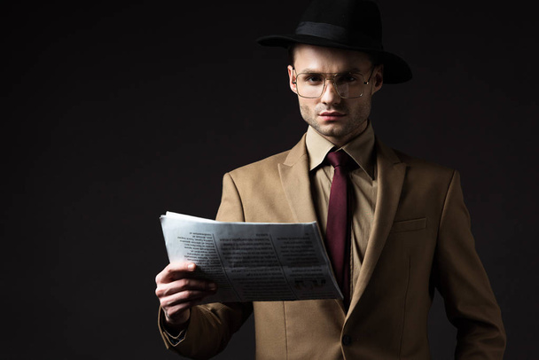 serious elegant man in beige suit, hat and eyeglasses holding newspaper isolated on black - Foto, Imagen