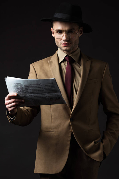 serious elegant man in beige suit, hat and eyeglasses holding newspaper isolated on black - Fotó, kép