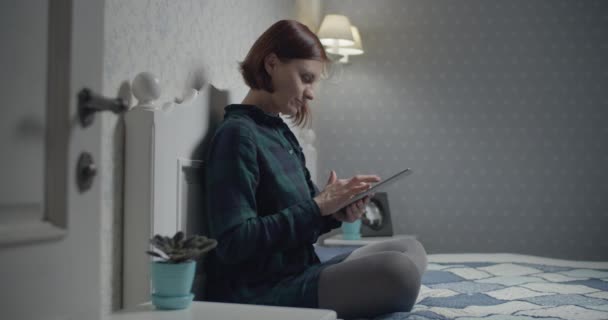Young female brunette adult working on tablet computer sitting on the bed. Woman touching gadget screen with fingers.  - Footage, Video