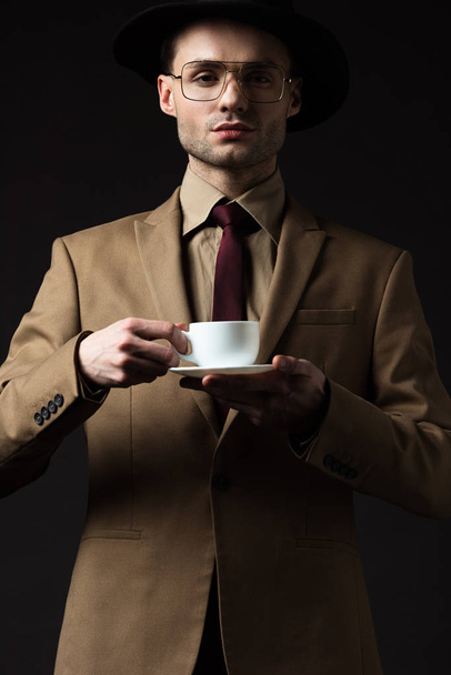 elegant man in beige suit holding coffee cup and saucer isolated on black - Photo, Image
