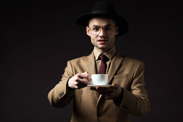 excited elegant man in beige suit, hat and eyeglasses holding coffee cup and saucer isolated on black - Fotó, kép