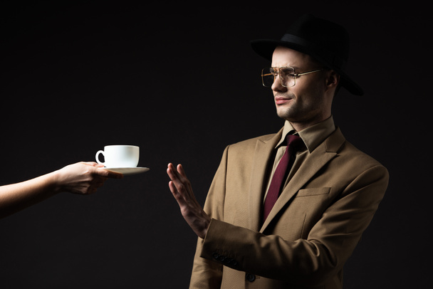 elegant man in beige suit, hat and eyeglasses refusing coffee from waiter isolated on black - Photo, Image