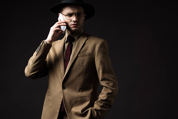 serious elegant man in beige suit, hat and eyeglasses talking on smartphone isolated on black - Photo, Image