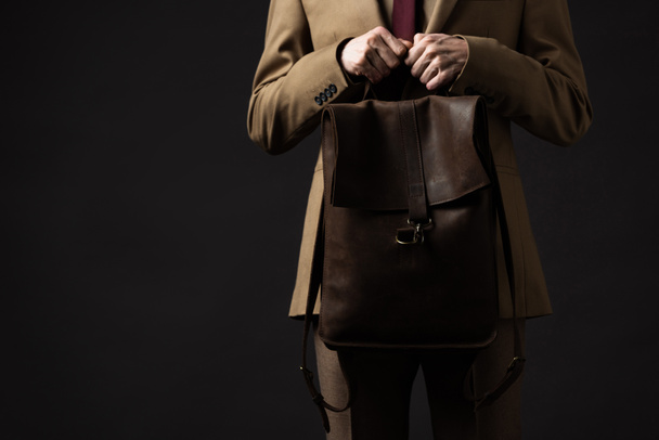 vue recadrée de l'homme élégant en costume beige tenant sac à dos en cuir brun isolé sur noir
 - Photo, image