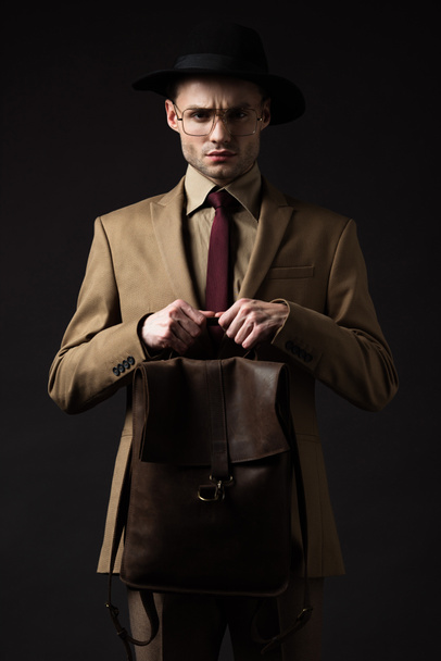 hombre elegante y tenso en traje beige, sombrero y gafas con mochila de cuero marrón aislada en negro
 - Foto, Imagen