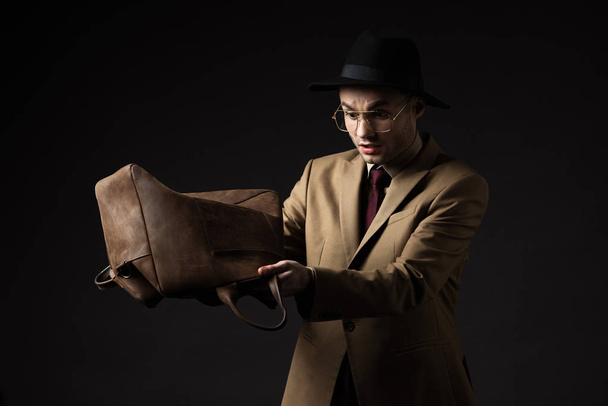 stressed elegant man in beige suit, hat and eyeglasses shaking out brown leather bag isolated on black - 写真・画像