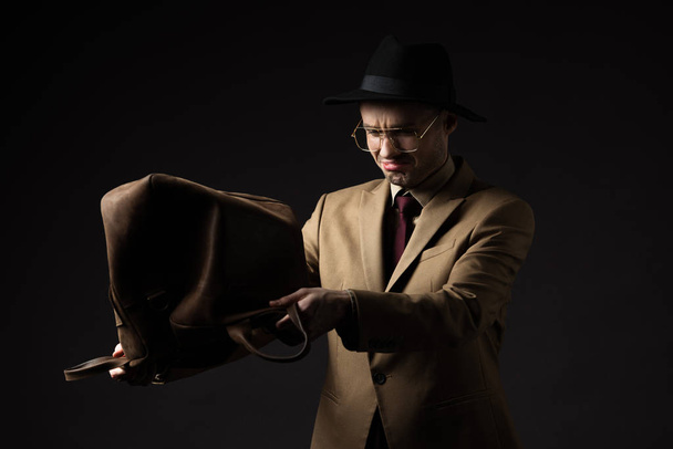 sad elegant man in beige suit, hat and eyeglasses shaking out brown leather bag isolated on black - Fotó, kép