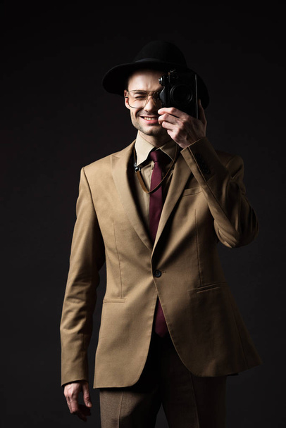 smiling elegant man in beige suit, hat and eyeglasses taking picture on film camera isolated on black - Zdjęcie, obraz