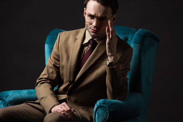 pensive elegant man in beige suit eyeglasses sitting in blue velour armchair isolated on black - 写真・画像