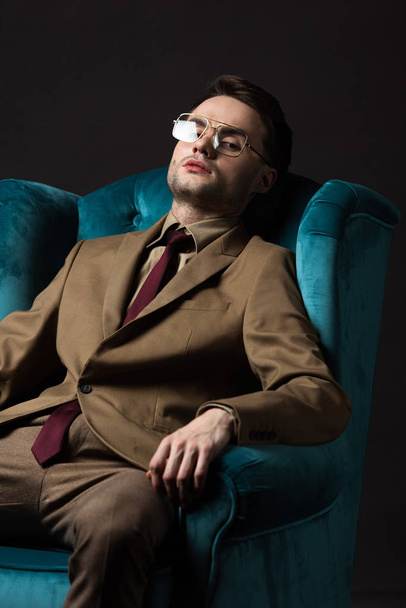 elegant man in beige suit eyeglasses sitting in blue velour armchair isolated on black - Foto, Imagen