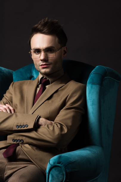 elegant man in beige suit eyeglasses sitting in blue velour armchair with crossed arms isolated on black - Foto, Imagem