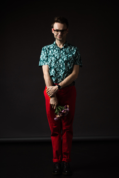 shy fashionable man in eyeglasses in blue colorful shirt and red pants holding floral bouquet on black - Foto, Bild