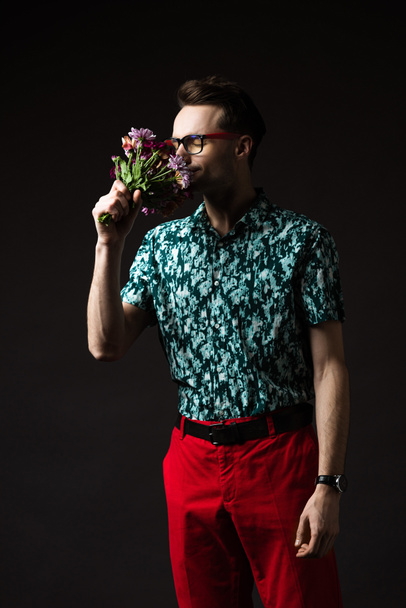 fashionable man in eyeglasses in blue colorful shirt and red pants smelling floral bouquet isolated on black - Foto, Imagen