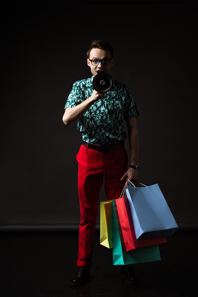modischer Mann mit Brille in blauem, buntem Hemd und roter Hose mit Einkaufstaschen schreit in Lautsprecher auf schwarz - Foto, Bild