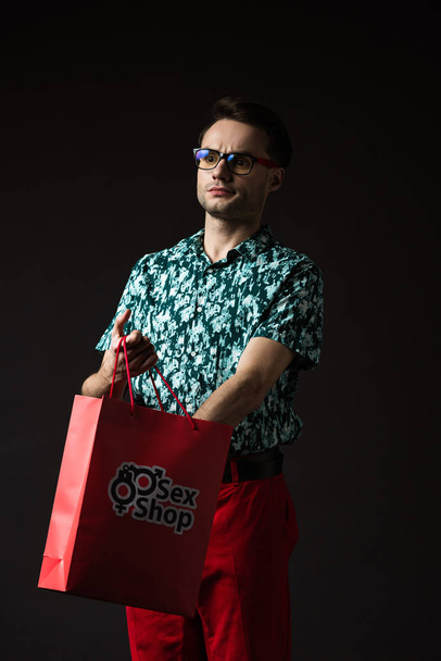 modieuze man in bril in blauw kleurrijk shirt en rode broek met rode boodschappentas uit sekswinkel geïsoleerd op zwart - Foto, afbeelding