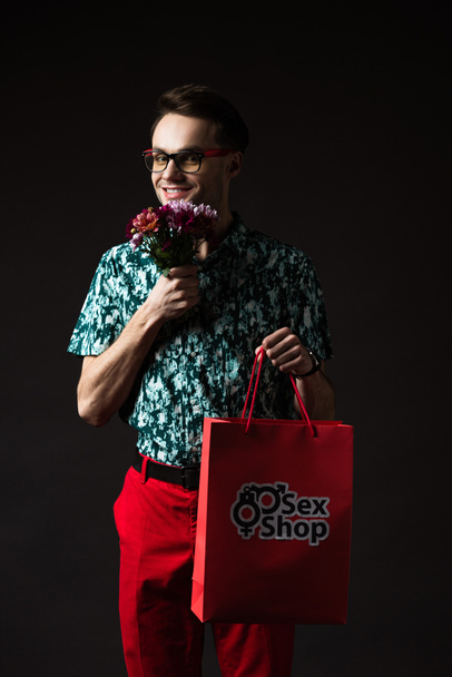 glimlachende modieuze man in bril in blauw kleurrijk shirt en rode broek met rode boodschappentas van seksshop en bloemen geïsoleerd op zwart - Foto, afbeelding
