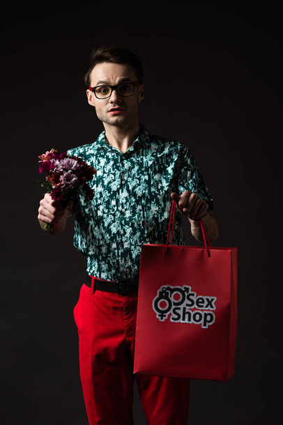 smiling fashionable man in eyeglasses in blue colorful shirt and red pants holding red shopping bag from sex shop and flowers isolated on black - Photo, Image