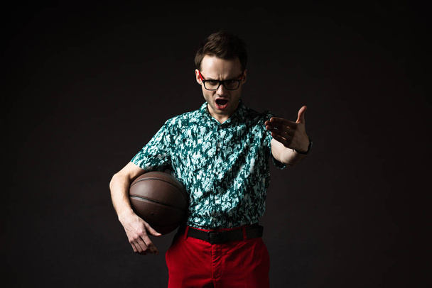 excited fashionable man in eyeglasses in blue colorful shirt and red pants holding basketball isolated on black - Foto, Bild
