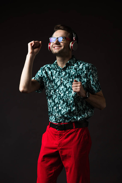 happy fashionable man in eyeglasses listening music in headphones and dancing isolated on black - Фото, зображення