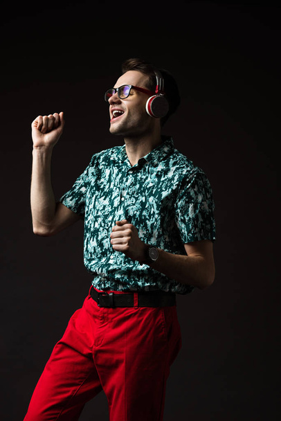 szczęśliwy modny mężczyzna w okularach słuchanie muzyki w słuchawkach i taniec odizolowany na czarny - Zdjęcie, obraz