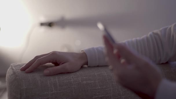 Stres moderního života: detailní záběr ženiných rukou nervózně poklepávajících prsty na pohovku, čekajících na zprávu na svém smartphonu v moderním prostředí měkkého světla, žádný slogan - Záběry, video