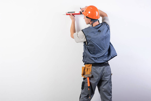 Builder with sealant. In work clothes and hard hat. On a light gray background. - Photo, Image