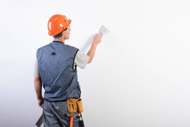 建築者はへらで壁を滑らかにします。作業服やハード帽子で。薄い灰色の背景で. - 写真・画像