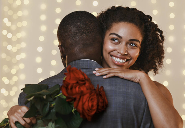Χαμογελώντας άφρο γυναίκα κρατώντας κόκκινα τριαντάφυλλα, ενώ αγκαλιάζει τον άντρα της - Φωτογραφία, εικόνα
