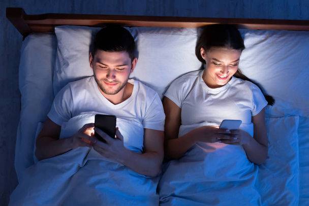 Couple Using Smartphones Lying In Bed At Home, Top View - Фото, изображение