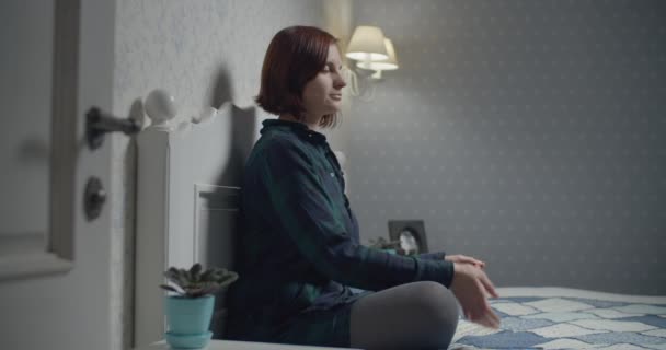 Young female seated in lotus position meditating with mobile application on the bed. Woman feeling tranquility while meditating in the evening.  - Záběry, video