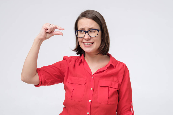 volwassen vrouw in bril en rood shirt tonen iets klein en klein met gebaar en kijken geschokt - Foto, afbeelding