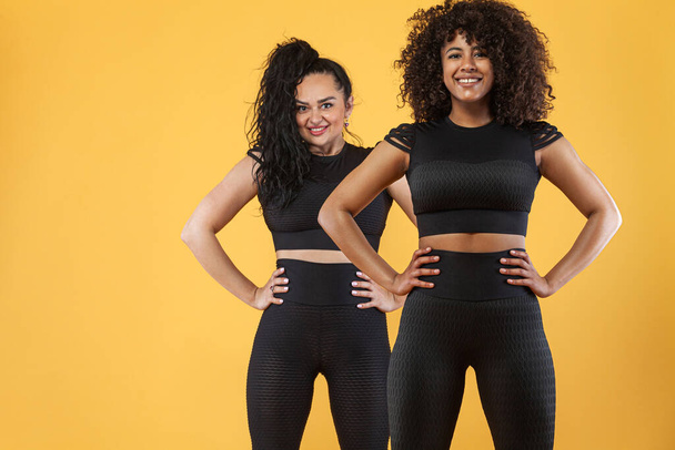 Two girls fitness trainer and size plus model - ward. Athletic women with black and white skin and curly hair, doing exercise on yellow background wearing sportswear. Fitness and sport motivation. - Foto, immagini